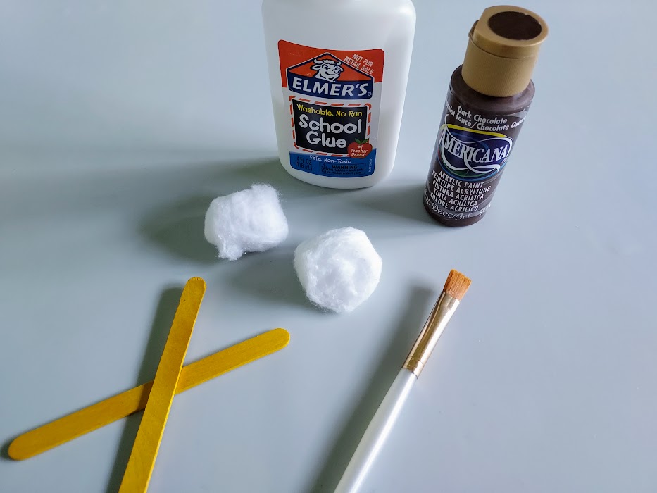 Two craft sticks, two cotton balls, a paintbrush, bottle of liquid glue, and bottle of brown paint setting out on table.