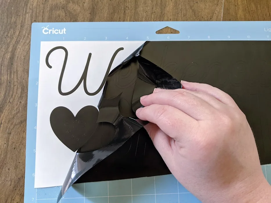White woman's hand peeling back excess black vinyl from design cut with Cricut machine.