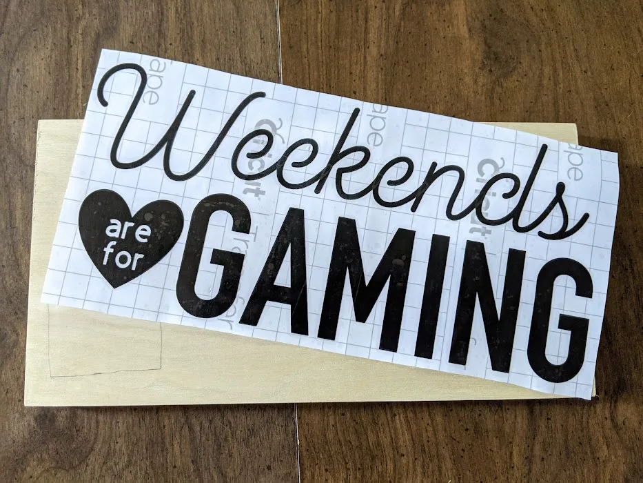 Black vinyl design with transfer tape over it. Design says Weekends are for (are for is inside a heart) Gaming. Design is laying at an angle over the top of a blank wooden sign.