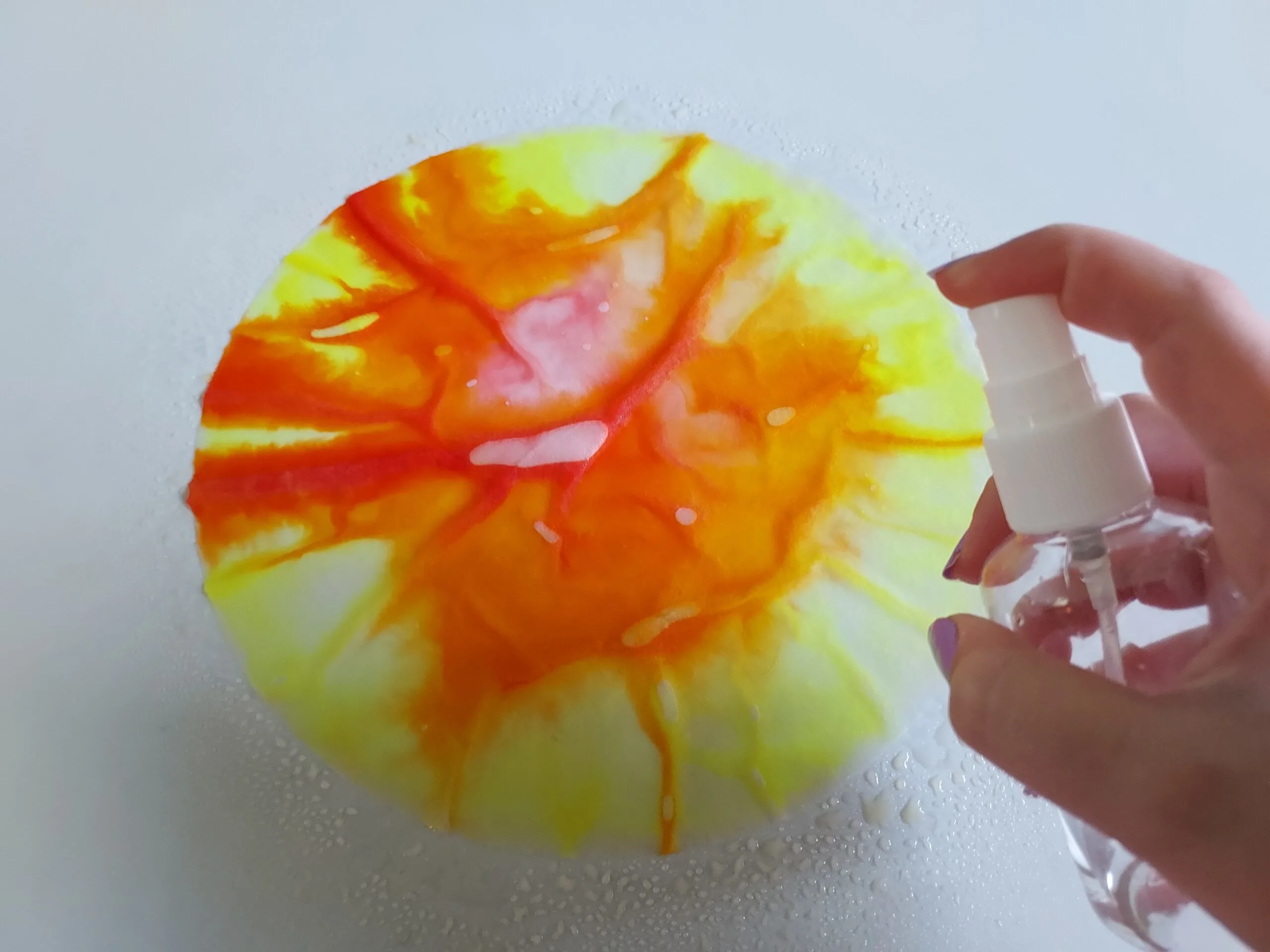 White person's hand using finger tip spray bottle to spray water on coffee filter colored with markers.