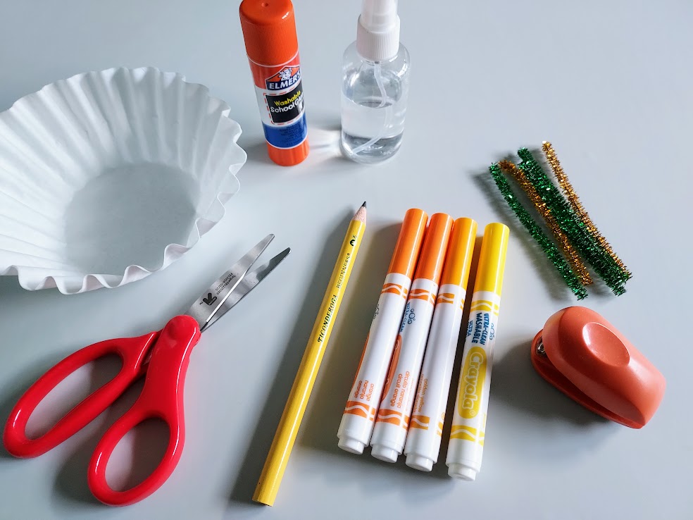 White coffee filters, small pair of red handled scissors, glue stick, finger tip spray bottle, pencil, orange and yellow washable markers, small stapler, and green and orange chenille stems spread out on workspace.