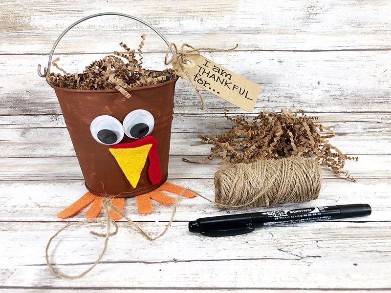 Turkey bucket filled with brown crinkle cut shredded paper and tag attached with twine. Tag says I am thankful for...