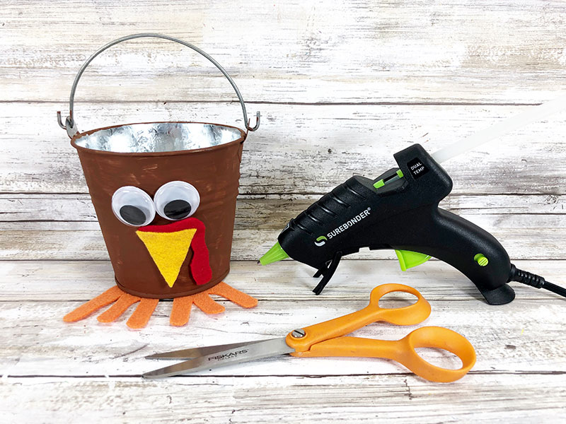 Brown metal bucket with turkey face and feet glued onto it. Turkey bucket next to scissors and hot glue gun.