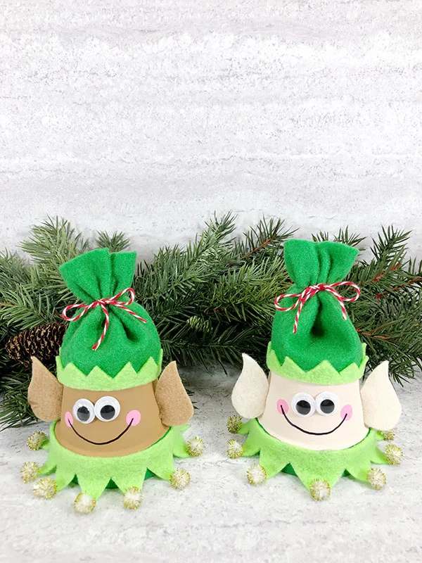 Two completed clay pot elf head ornaments sitting next to each other in front of evergreen garland. Both elves have green felt hats and collars. One has lighter skin tone and one has darker skin tone.