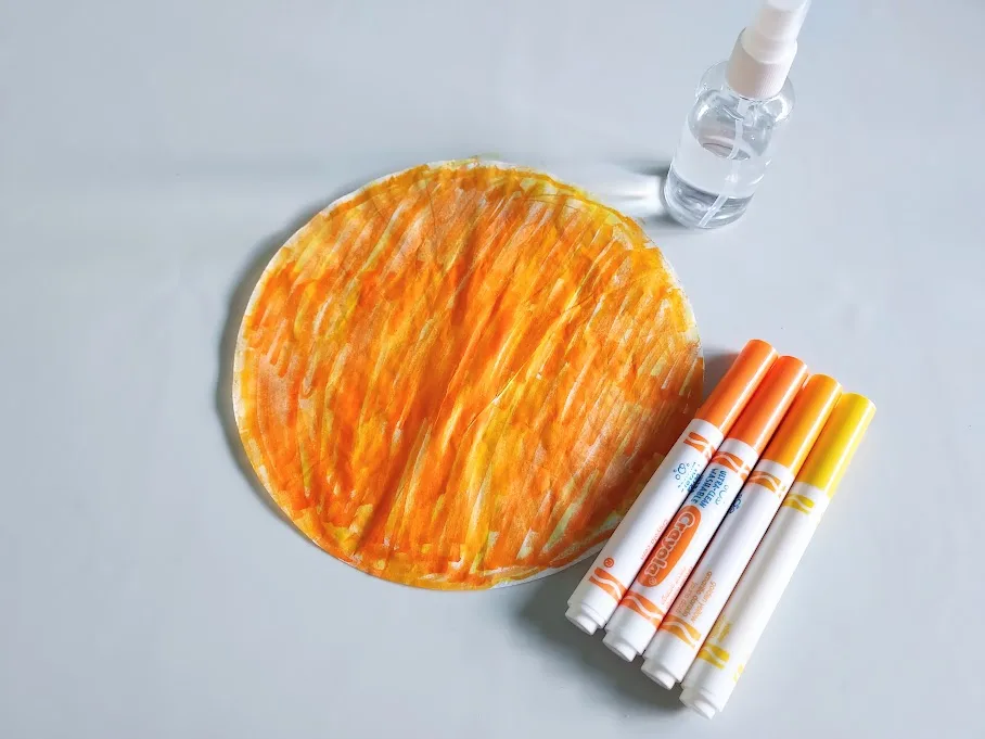 Round coffee filter colored with orange and yellow washable markers. Markers laying next to filter and small spray bottle filled with water.