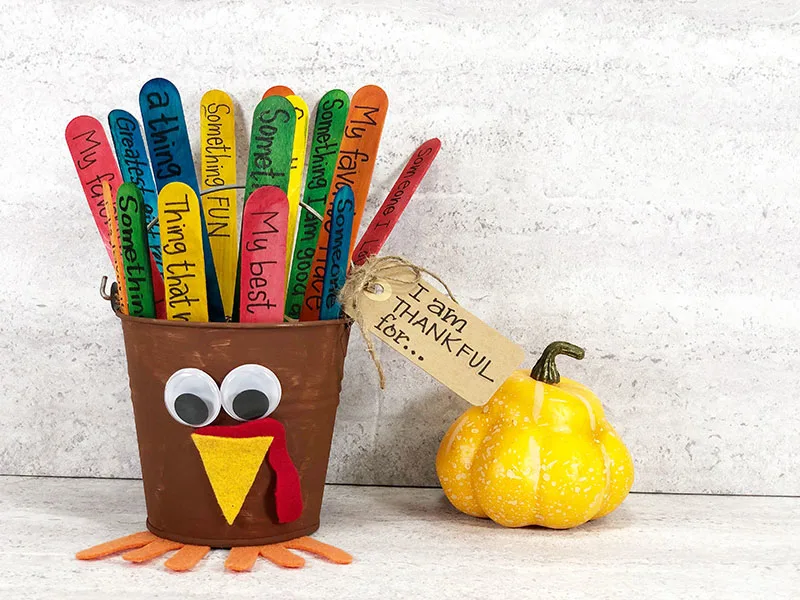 Completed thankful turkey bucket craft with gratitude prompts written on craft stick tail feathers in the bucket. Turkey is sitting next to a small yellow gourd decoration.