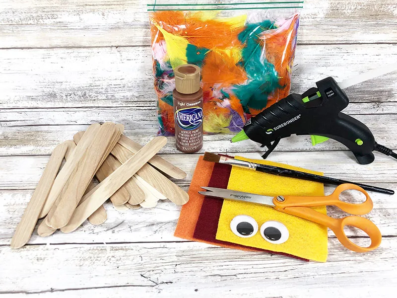 Craft sticks, felt, googly eyes, scissors, brown paint, craft feathers, and hot glue gun on work surface.