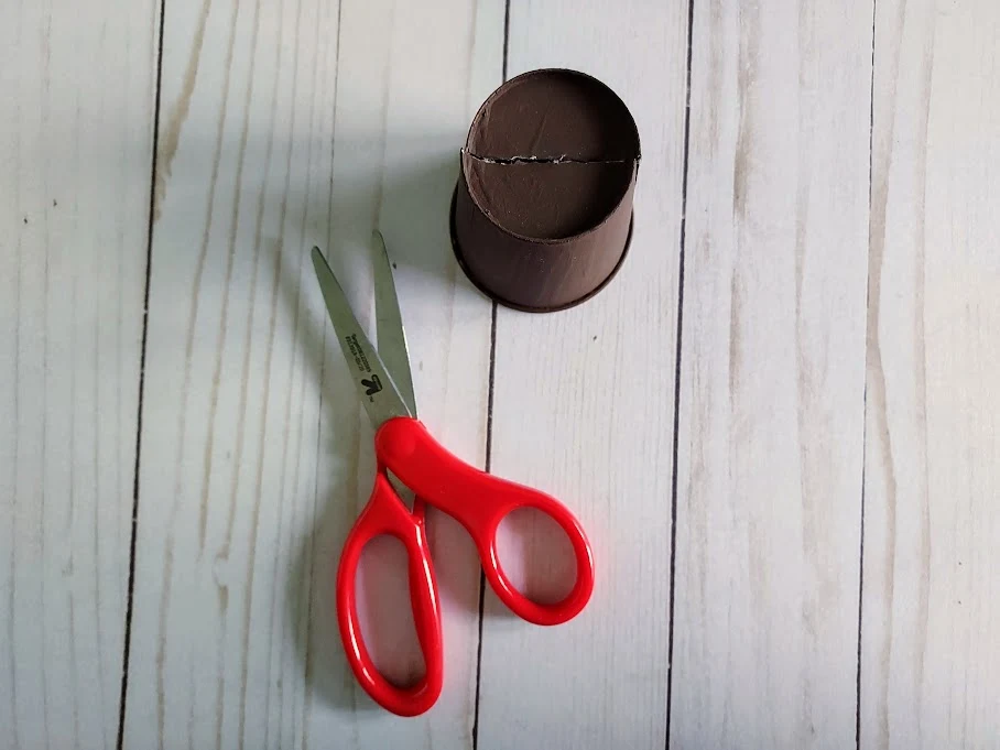 Overhead view of upside down brown painted paper cup with line cut across bottom of cup. Pair of scissors next to cup.