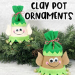 Black text at top reads Elf Clay Pot Ornaments. Black text at bottom reads Christmas Craft for Kids. Two finished elf clay pot crafts arranged by a garland of evergreen. One elf is painted with lighter skin tone and one is painted with darker skin tone. They are wearing green felt hats.