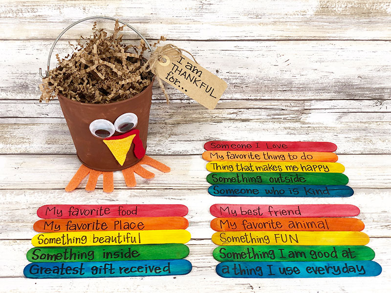 Turkey bucket filled with crinkle paper sitting next to colored jumbo craft sticks with gratitude prompts written on them in black marker.