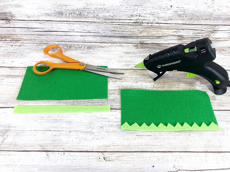 Demonstrating how elf hat with trim should look with dark green felt cut in larger rectangle and thin strip of light green felt cut out with one jagged side.