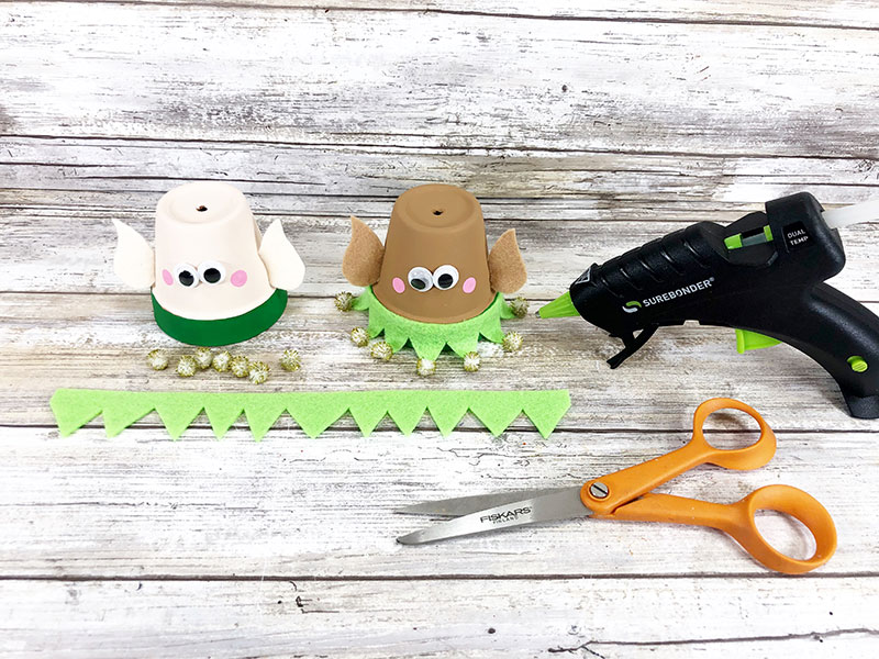 Light green felt strip cut with one jagged side laying in front of both clay pot elf heads. Clay pot on the right has green felt glued around the bottom like a collar and gold pom poms glued to the points.