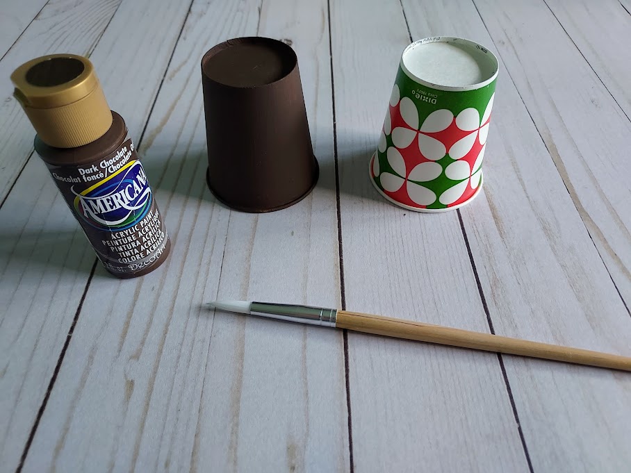 Brown painted paper cup set upside down next to unpainted cup. Bottle of brown paint and paint brush near cups.