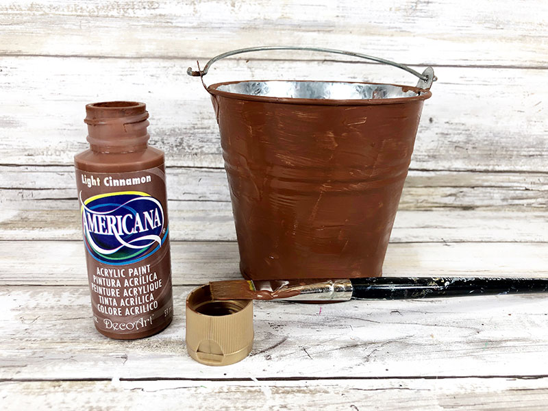 Small metal bucket painted brown on the outside. Bottle of brown acrylic paint and paint brush nearby.