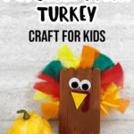 Black text at top says Popsicle Stick Turkey Craft for Kids above completed craft project of turkey made out of popsicle sticks, craft feathers and felt. Turkey sitting next to yellow gourd decoration.