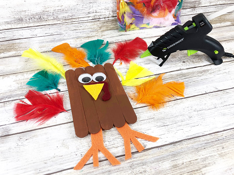 Brown painted craft sticks with googly eyes, beak and snood glued on. Orange felt feet positioned at bottom of turkey and craft feathers arranged around it on the table next to glue gun.