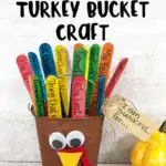 Thankful Turkey Bucket Craft in black text at top of picture of completed craft project. Small metal bucket painted brown with googly eyes and felt beak and feet glued to it. Multiple colors of jumbo craft sticks sticking out of it to look like tail feathers with things written on them. Brown kraft tag hanging off handle says I am thankful for...