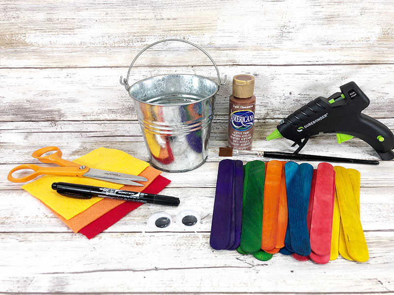 Yellow, orange, and red felt, scissors, black permanent marker, two googly eyes, assorted colored jumbo craft sticks, small plain metal bucket with handle, brown paint, paint brush, and hot glue gun laying out for project.