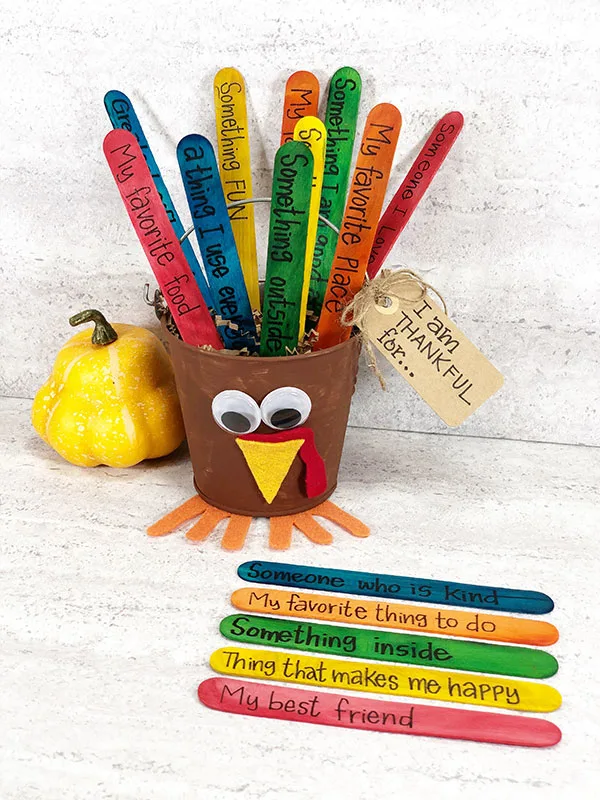 Small metal bucket painted and made to look like a turkey with craft stick tail feathers sticking out of it. Some colored craft sticks laying neatly on table by turkey bucket showing thankful prompts such as someone who is kind and my favorite thing to do. Small yellow gourd decoration sitting to the left of the turkey bucket.