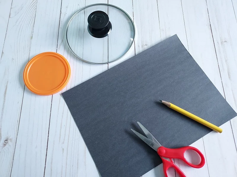 Glass pot lid and smaller round orange lid next to black construction paper, scissors, and pencil.