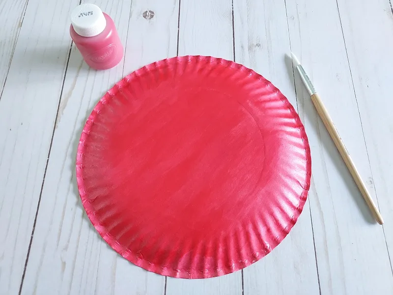 Back of paper plate painted completed red. Paint brush and bottle of washable red paint next to it.