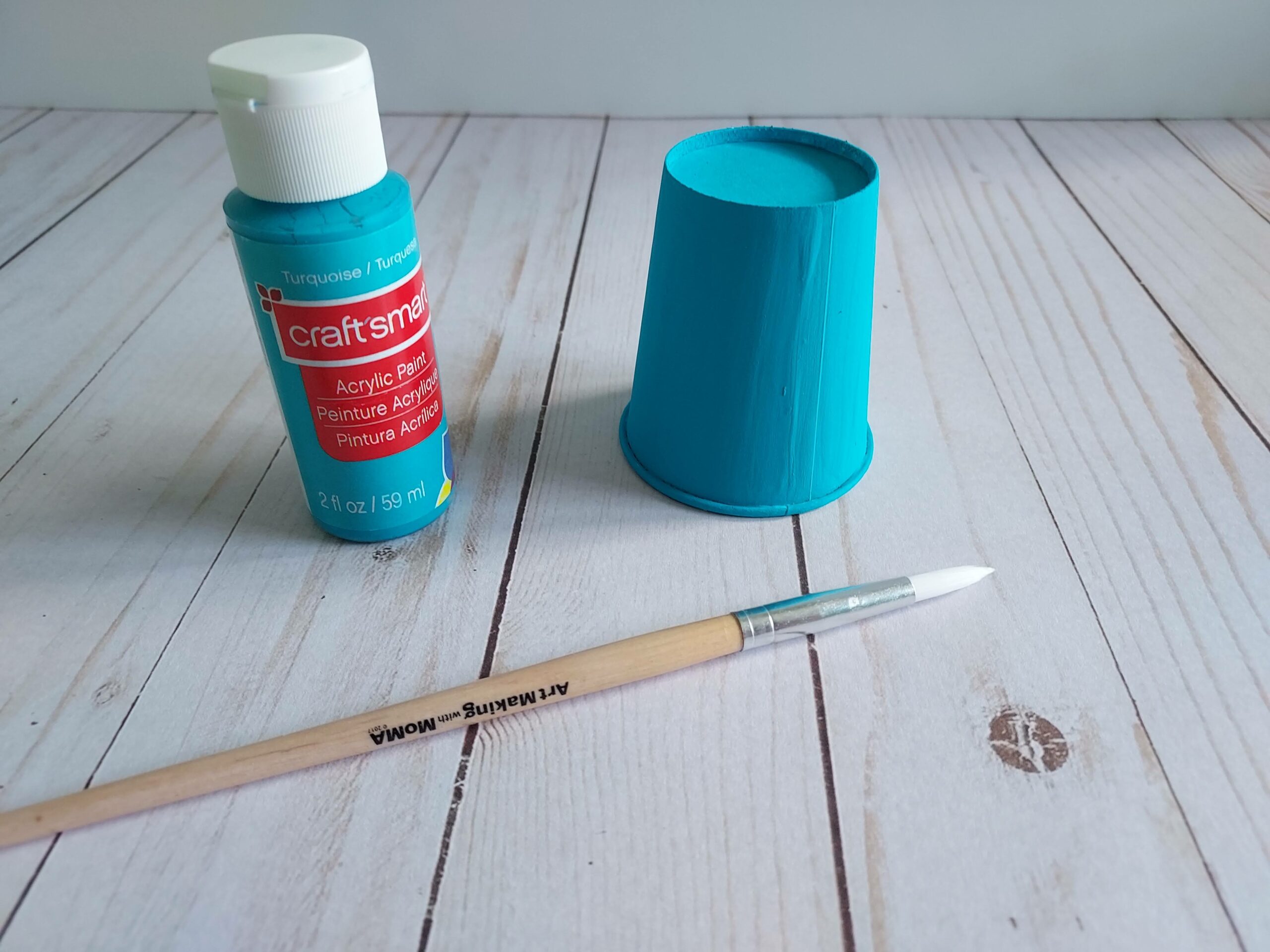 Blue paint bottle and paint brush next to blue painted paper cup.