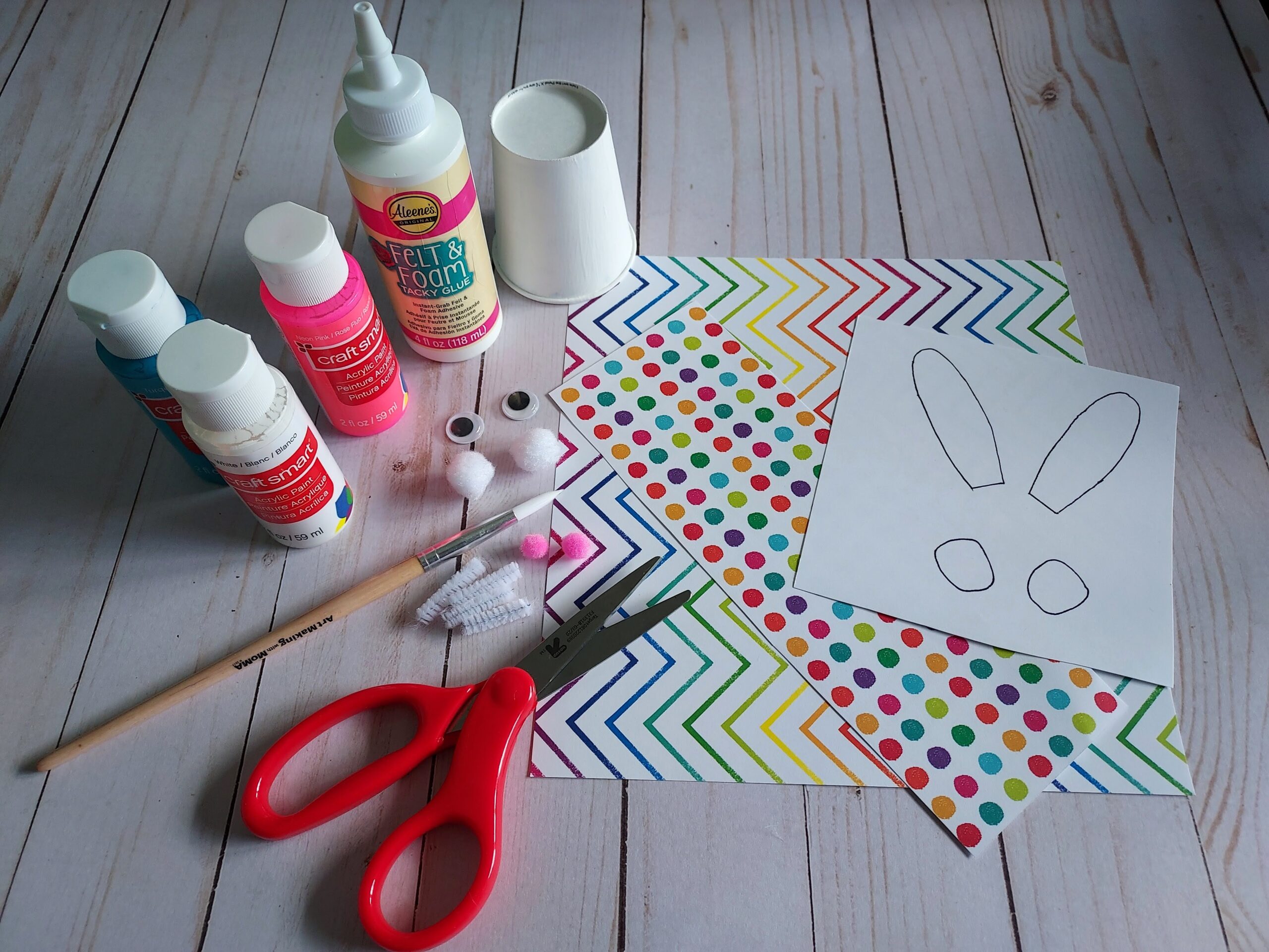 Craft supplies for making paper cup bunnies gathered on table: paint, cup, tacky glue, scissors, patterned cardstock paper, googly eyes, pom poms, chenille stem, and printable template for ears and feet.