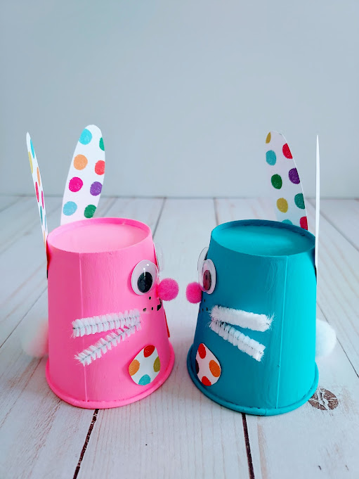 Side view of two finished paper cup bunnies that are facing each other to show their pom pom tails on the back. Left bunny is pink and right one is blue.
