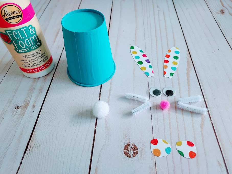 Tacky glue and blue painted cup next to googly eyes, chenille stem, pom poms, and cut out patterned cardstock laying out to make a bunny face.