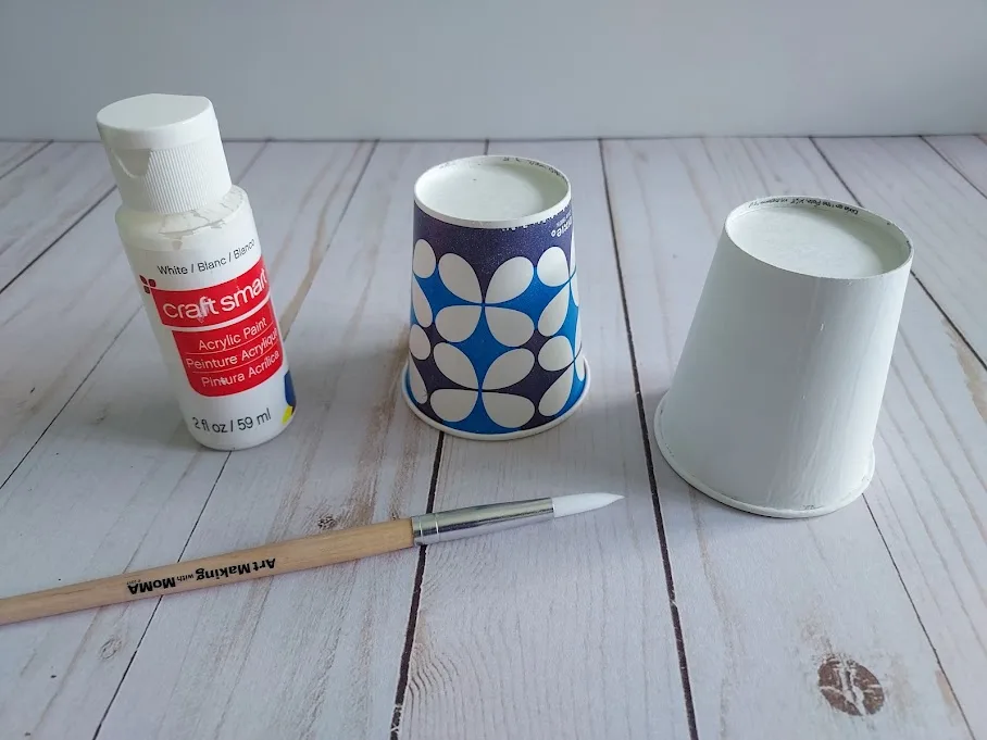 Unpainted patterned Dixie paper cup sitting next to one painted over with white paint.