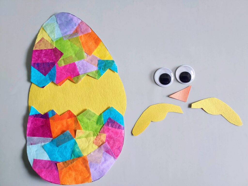Yellow circle body for baby chick glued behind hatching egg pieces. Paper wings, beak, and googly eyes laying on table next to craft project waiting to be added.