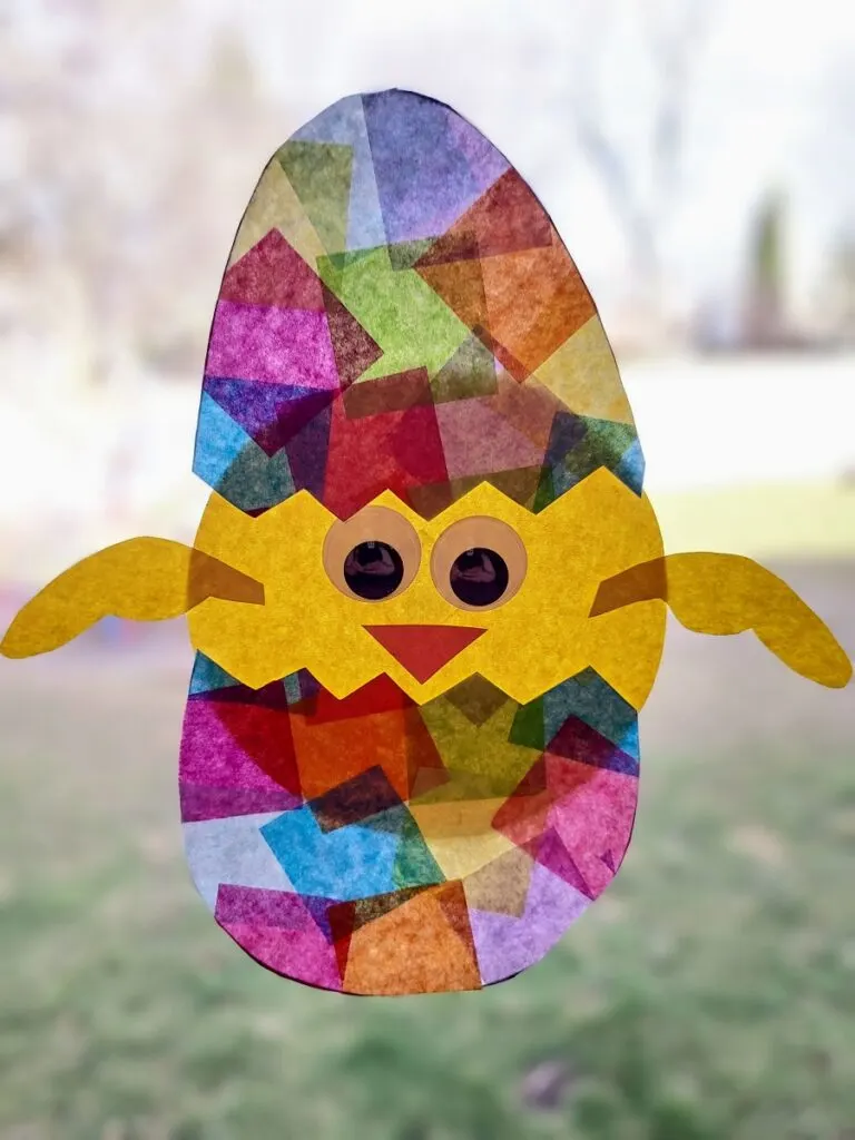 Hatching baby chick suncatcher craft made with tissue paper and construction paper hanging in sunny window.