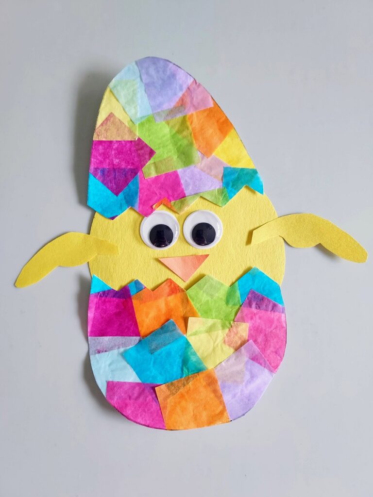 Completed hatching chick suncatcher craft. Egg is made with colorful tissue paper glued to white printer paper. Egg is cut in half in a zig zag pattern. Baby chicken made with construction paper and googly eyes is glued between opening of egg halves.