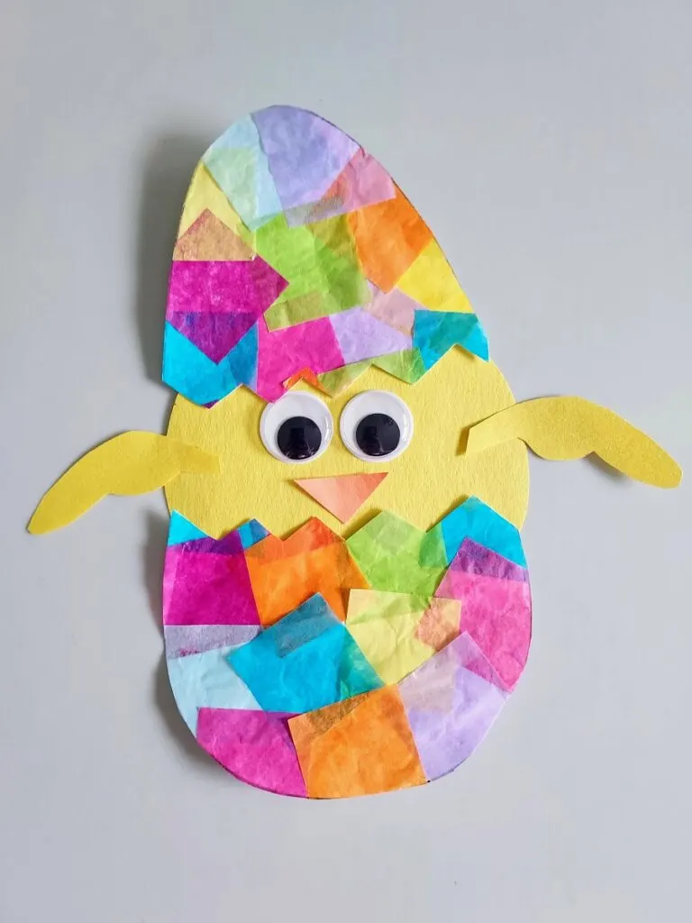 Completed hatching chick suncatcher craft. Egg is made with colorful tissue paper glued to white printer paper. Egg is cut in half in a zig zag pattern. Baby chicken made with construction paper and googly eyes is glued between opening of egg halves.