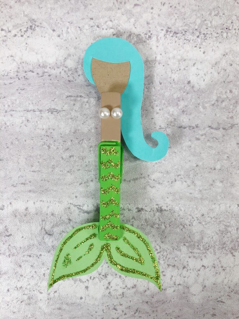 Close view of mermaid craft made with paper and wooden clothespin. Mermaid has aqua hair, light brown skin tone, pearl swim top, and lime green tail decorated with glitter glue.