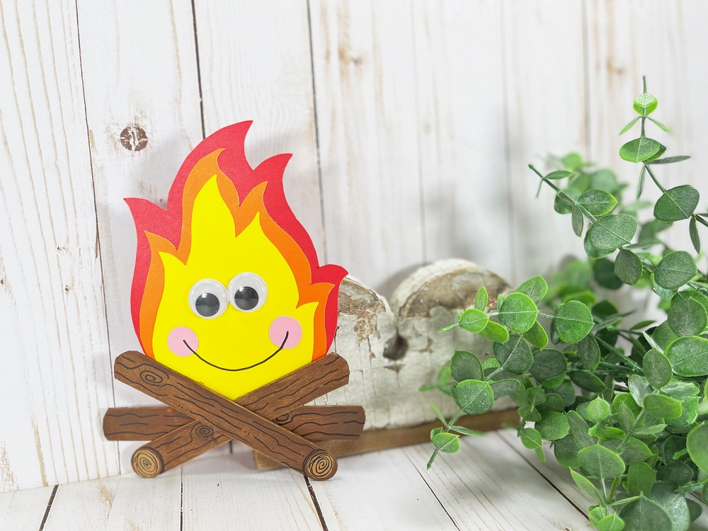 Cute paper and popsicle stick campfire craft propped up and leaning against something decorative next to a fake plant.