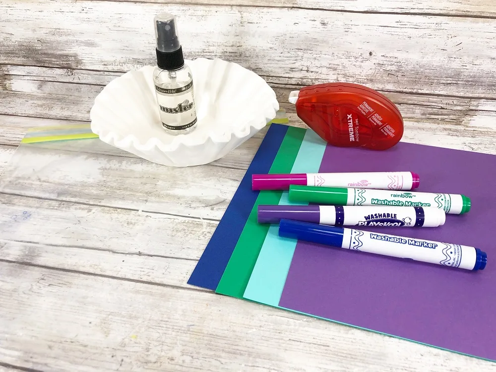 Materials for making a coffee filter mermaid tail laying out. Includes coffee filters, small spray bottle, cardstock paper in blue, green, and purple, assorted washable markers, and adhesive.