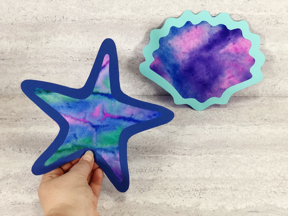 Left foreground is a hand holding a completed coffee filter starfish suncatcher. In the right background is a completed coffee filter seashell suncatcher.