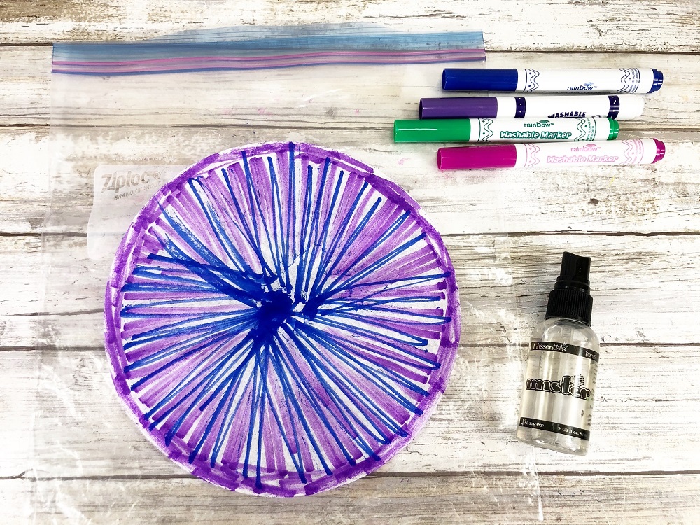 Overhead view of coffee filter colored with purple, pink, and blue markers. Coffee filter is on top of a plastic zip top bag. Markers and small spray bottle filled with water lay next to it.