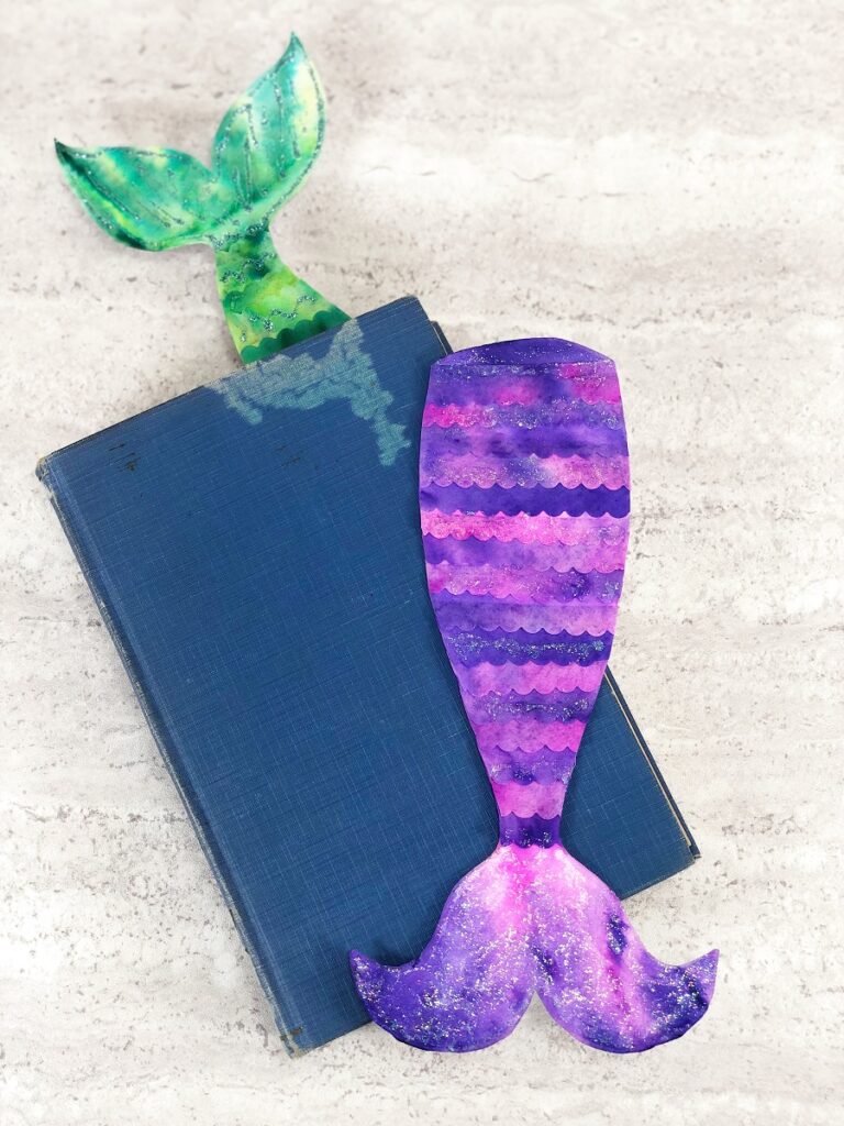 Top down view of two completed coffee filter mermaid bookmarks. Green tie dyed one sticking out of closed book. Purple and pink tie dyed mermaid tail is laying on top of blue hardcover book.