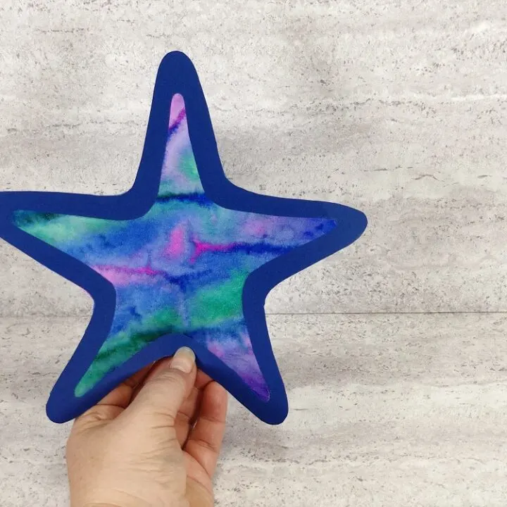 Hand holding finished starfish craft for kids made with a tie dyed coffee filter and dark blue cardstock frame outline.