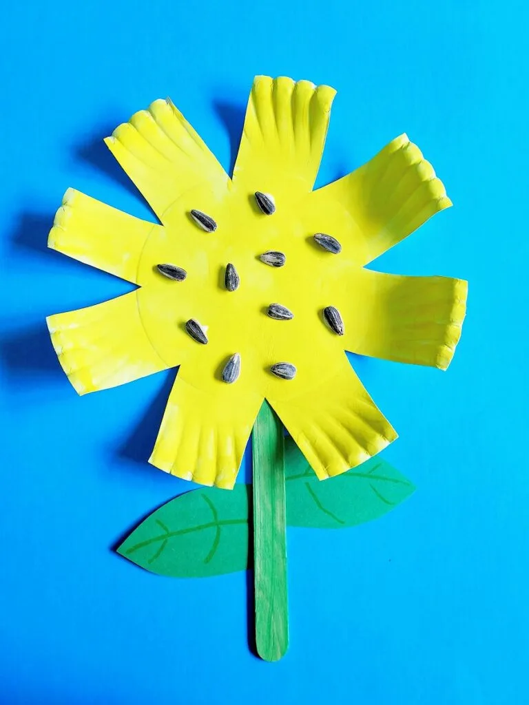 Completed paper plate sunflower craft made with a paper plate cut like a flower, painted yellow, and sunflower seeds glued in the middle. Popsicle stick painted green glued to plate. Green paper leaves cut out and glued to stick. Completed craft laying on a blue background.