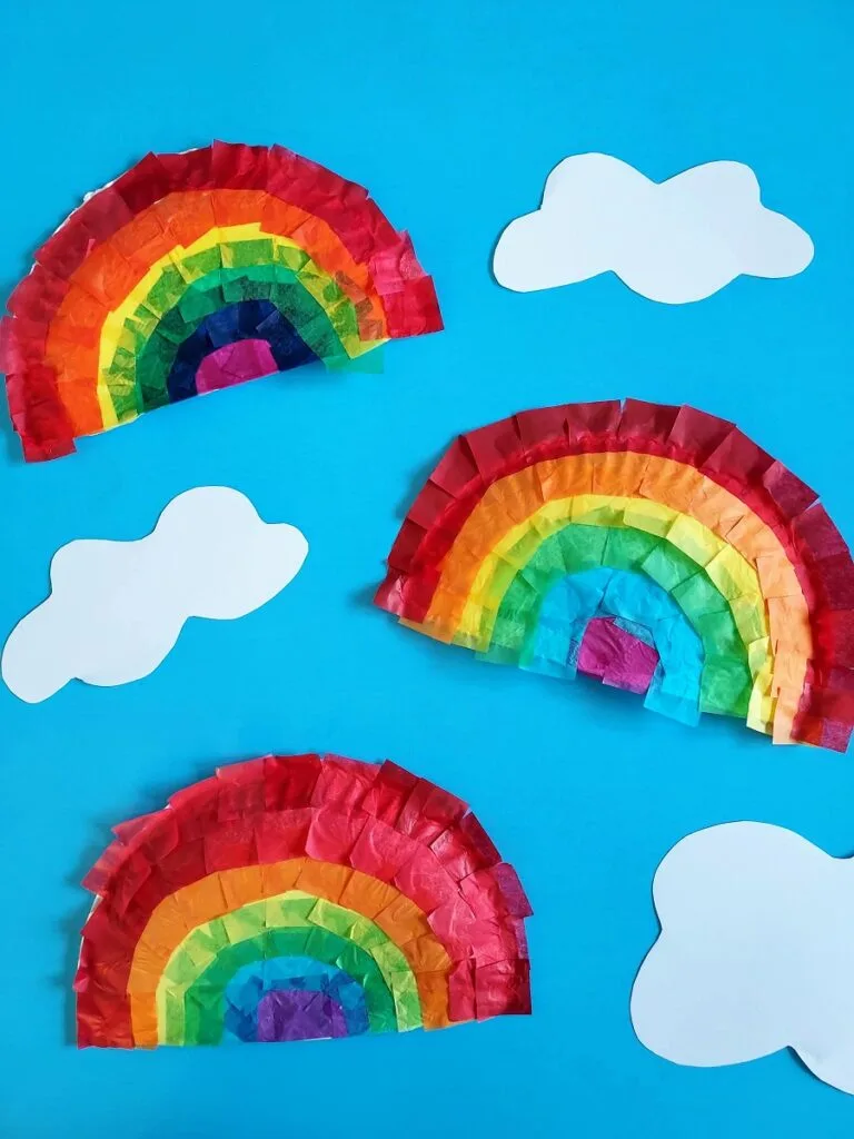 Three finished paper plate tissue paper rainbows arranged on sky blue paper around white clouds cut out from paper.