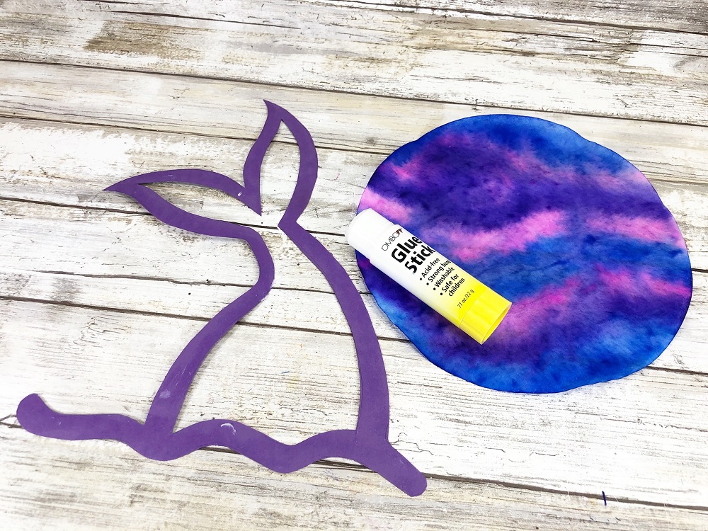 Mermaid tail suncatcher frame cut out of purple cardstock laying next to glue stick and colorful coffee filter.