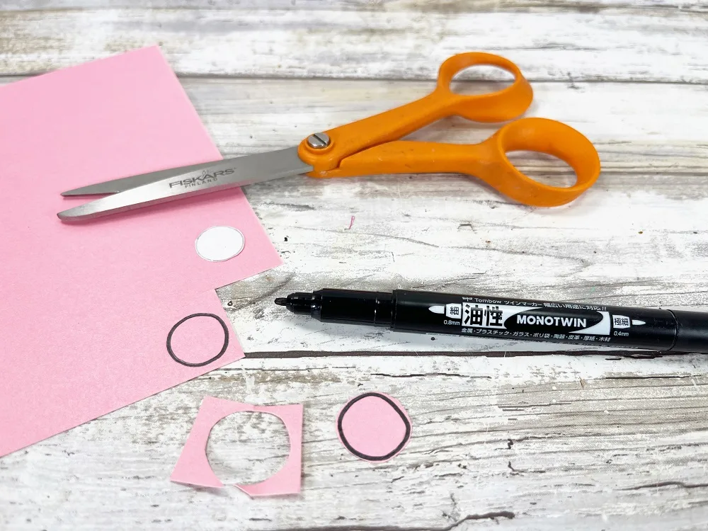 Two small pink circles cut out of the pink cardstock paper. Scissors, black marker and rest of pink paper laying there.