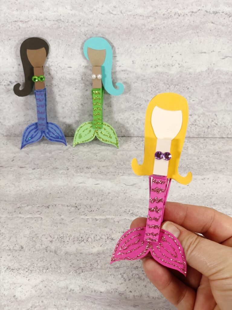 Hand holding clothespin mermaid with yellow hair, light skin tone, and pink tail in foreground. Two more clothespin mermaids with dark skin tones are in the background.