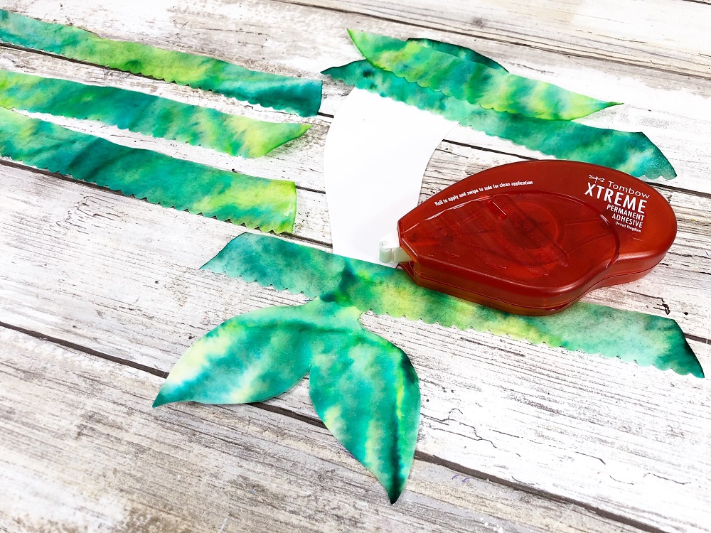 Attaching green tie dye coffee filter strips onto mermaid tail bookmark using an adhesive runner aka glue tape.