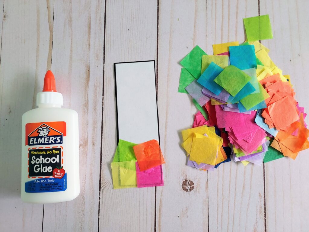 Bottle of Elmer's white school glue laying next to single cutout bookmark template with a few pieces of tissue paper glued to it.