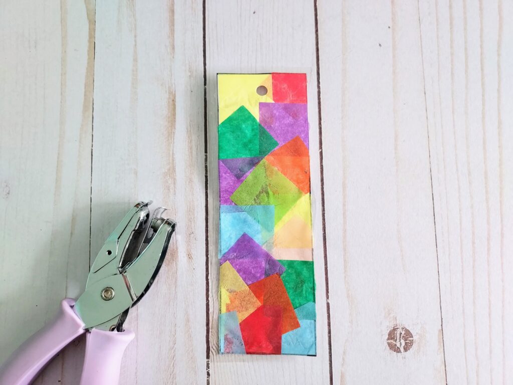 Hand hole puncher laying next to laminated tissue paper bookmark with hole near the top.