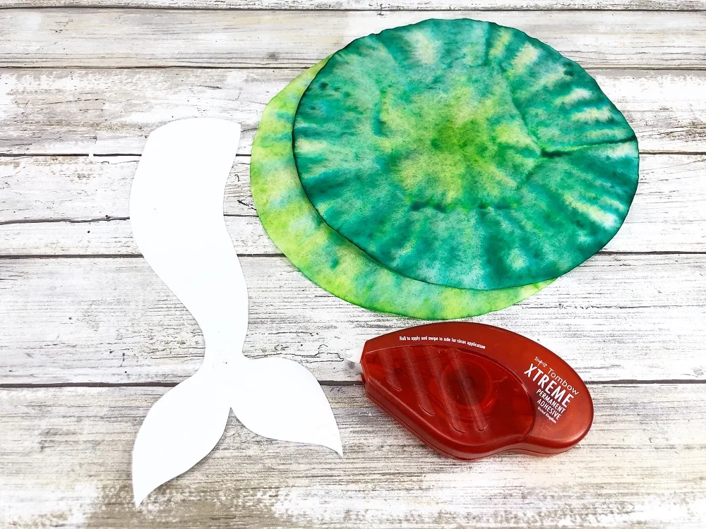 Mermaid tail craft template cut out next to green dyed coffee filters and a red adhesive runner.
